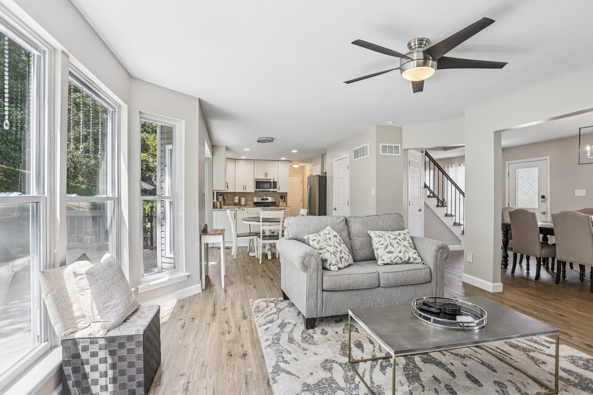 Bright, airy open-plan living room with gray sofa, glass coffee table, patterned rug, kitchen and dining area, large windows, and ceiling fan.