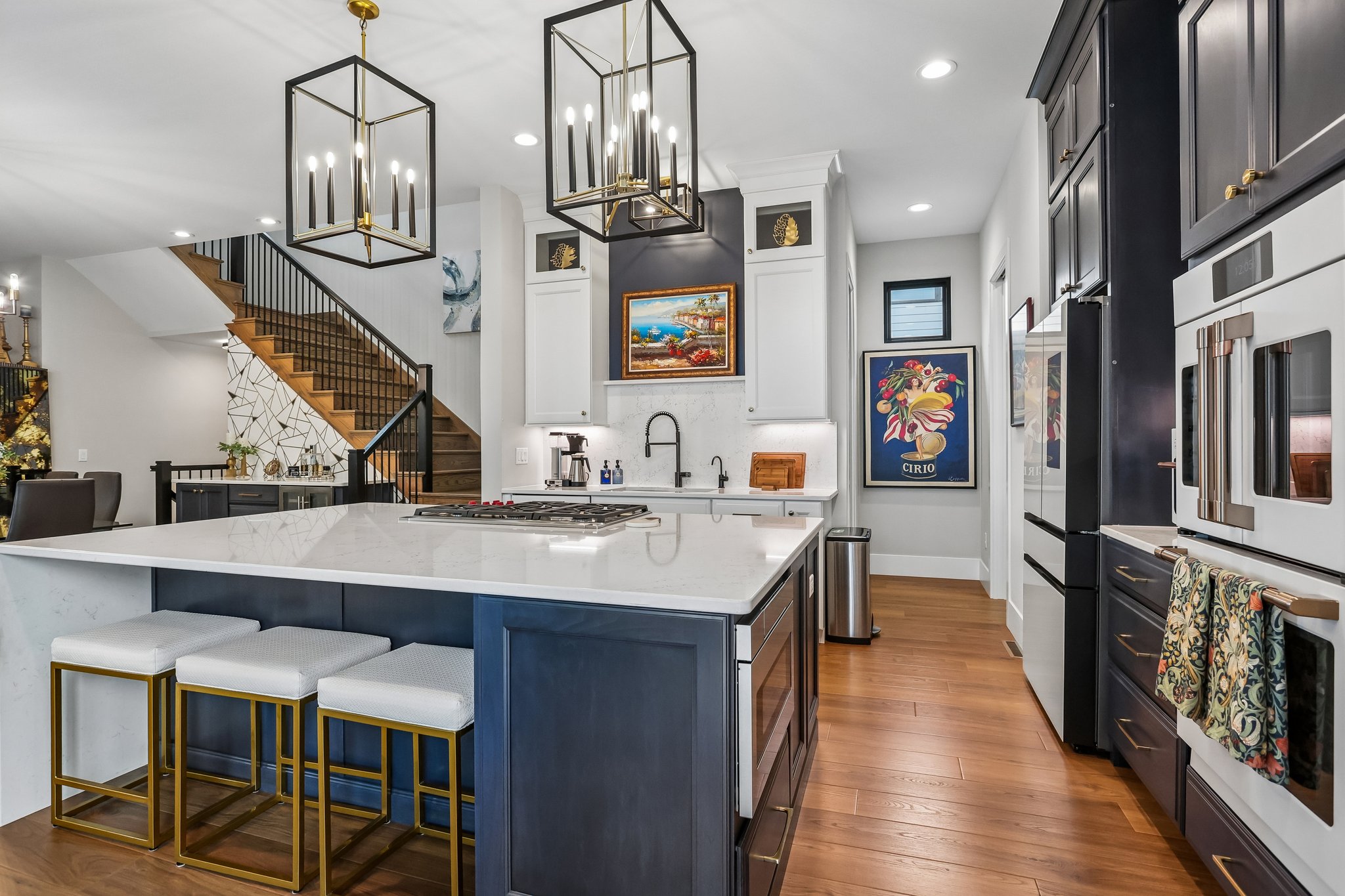 Modern kitchen with a large island, sleek black and white cabinetry, decorative lighting, and an inviting wooden staircase in the background.