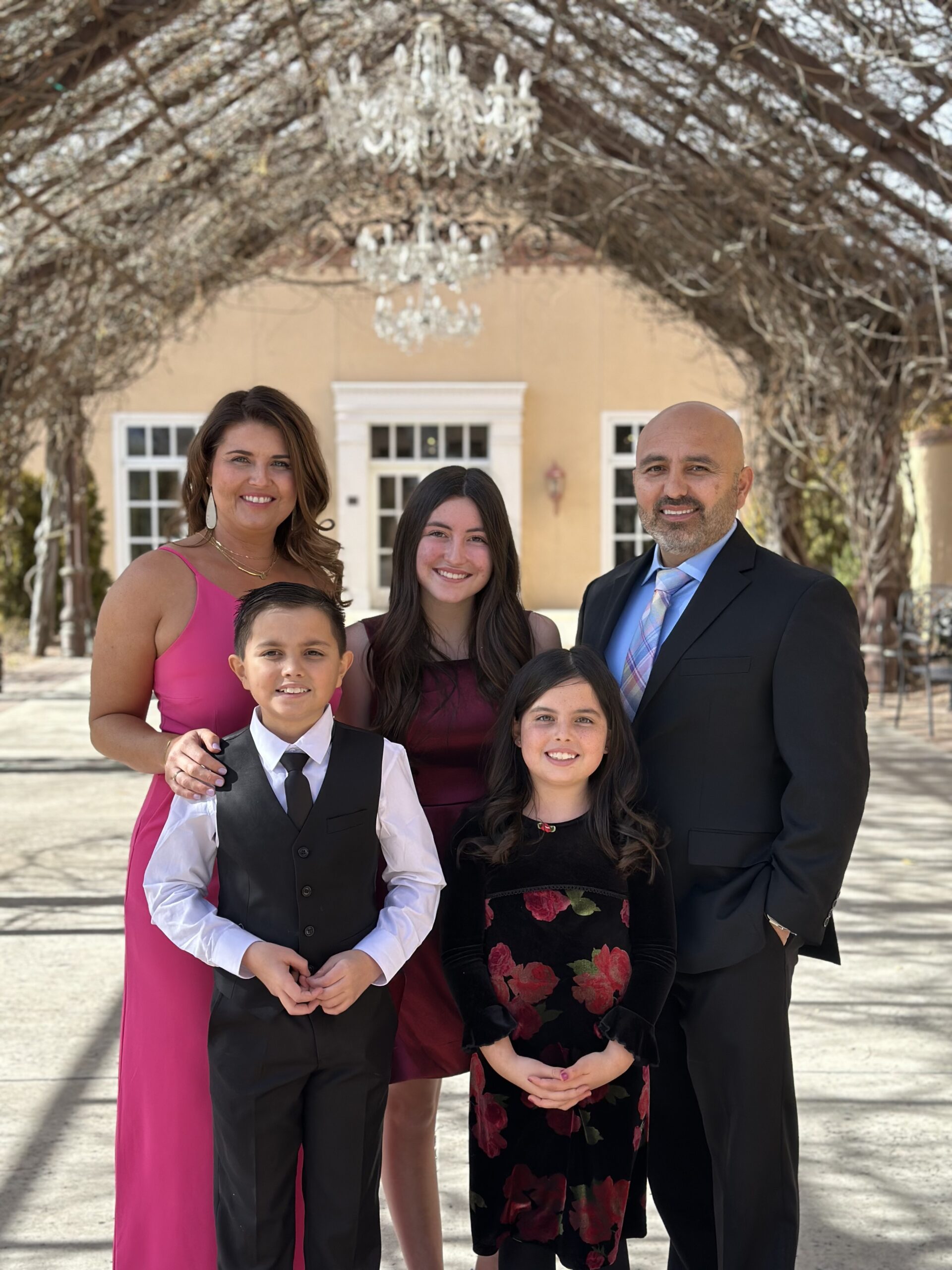 A family poses outdoors under a vine-covered pergola, dressed in formal attire: a woman in pink, a man in a suit, and three children in elegant outfits.